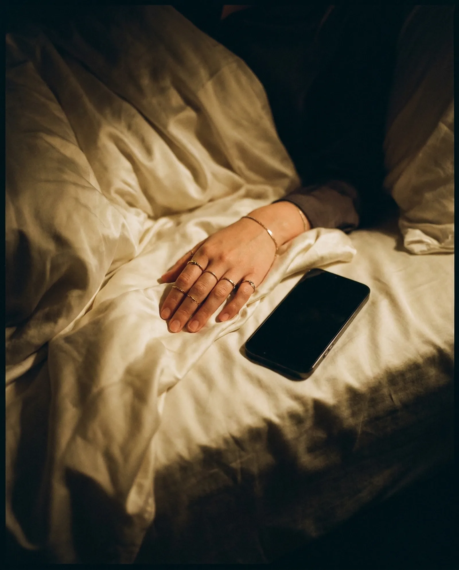 A hand with fine gold rings resting on ivory silk beside a phone